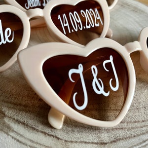 May include: A pair of heart-shaped sunglasses with brown lenses and a white inscription that reads "J&J". The sunglasses are made of a light pink plastic.