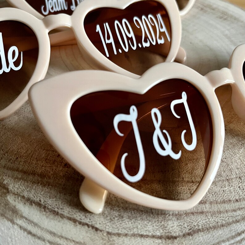 May include: A pair of heart-shaped sunglasses with brown lenses and a white inscription that reads "J&J". The sunglasses are made of a light pink plastic.