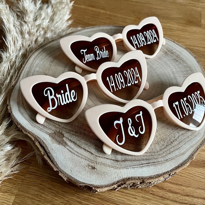 May include: Set of five pink heart-shaped sunglasses with white text. The sunglasses say "Bride", "Team Bride", "J&J", and two dates.