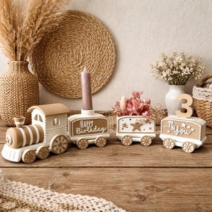 May include: A decorative wooden train set featuring a locomotive, two cargo cars, and a caboose. The train is painted in white and tan. The cars display the text Happy Birthday and To You, with a wooden number 3 and a pink candle holder.