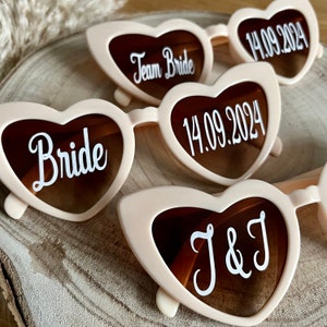 May include: Set of four heart-shaped sunglasses with white text. The sunglasses are pink with brown lenses. The text on the sunglasses reads "Bride", "Team Bride", "14.09.2024", and "J&J".