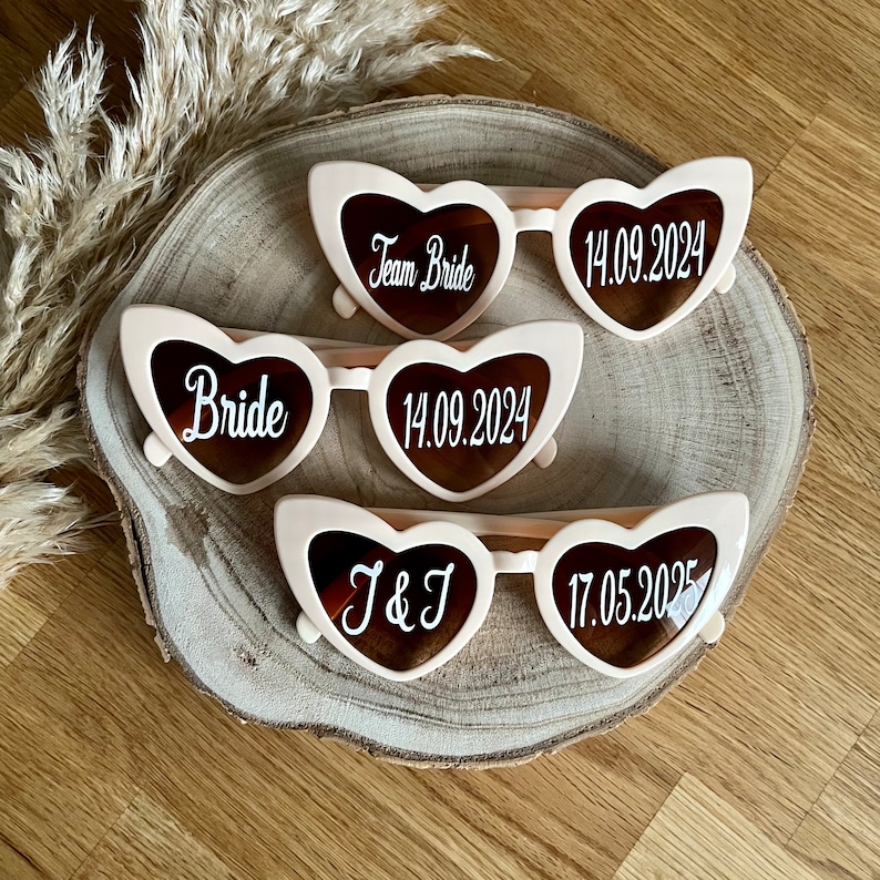 May include: Three pairs of heart-shaped sunglasses with different inscriptions. The first pair says "Team Bride", the second pair says "Bride 14.09.2024", and the third pair says "J & J 17.05.2025".