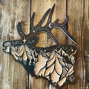 May include: A black and white silhouette of an elk's head with a mountain range and starry sky inside. The elk's antlers are curved and point upwards. The mountain range is detailed with peaks and valleys. The starry sky is filled with small white stars.