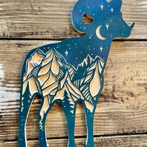 Può includere: Silhouette di montone in legno con un design a strati. Il corpo del montone presenta una catena montuosa e una scena di foresta nei toni naturali del legno. Il cielo è blu scuro con una luna crescente e stelle. Il montone è in piedi su quattro zampe.