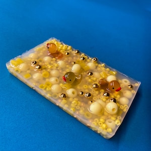 Könnte beinhalten: Ein rechteckiger, transparenter Seifenblock mit gelben und goldenen Perlen und drei gelben Gummienenten. Die Enten haben rote Schnäbel und sind teilweise in die Seife eingetaucht.