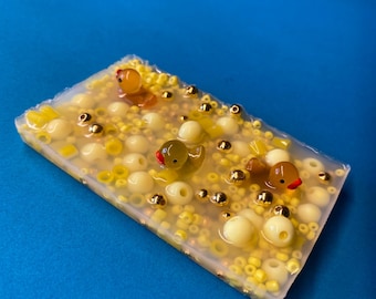 Könnte beinhalten: Ein rechteckiger, transparenter Seifenblock mit gelben und goldenen Perlen und drei gelben Gummienenten. Die Enten haben rote Schnäbel und sind teilweise in die Seife eingetaucht.
