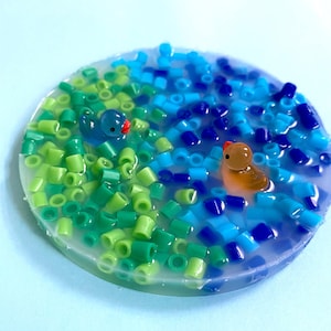 Könnte beinhalten: Ein runder, transparenter Harz-Untersetzer mit grünen und blauen Perlen, die in das Harz eingebettet sind. Zwei kleine Glasfiguren von Enten sitzen im Harz.