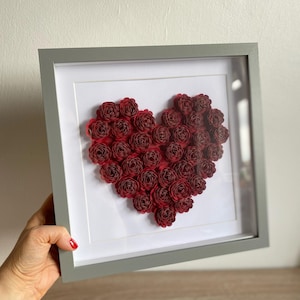 May include: A framed artwork featuring a heart shape made of many deep red paper roses. The heart is on a white background within a grey frame. The artwork is held by a person.