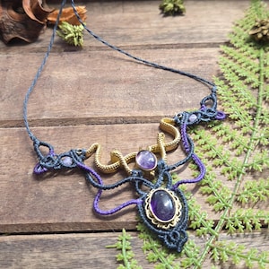 Könnte beinhalten: Eine Makramee-Halskette aus dunkelgrauer und violetter Kordel, die in einem komplexen Muster gewebt ist. Das Design enthält ein goldfarbenes Metallelement in Schlangenform und zwei ovale, violette Amethyst-Edelsteine in goldfarbenen Fassungen.