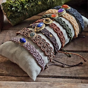 Könnte beinhalten: Eine Sammlung von sieben Makramee-Armbändern mit geflochtenen Kordelmustern in verschiedenen Farben, darunter Lavendel, Braun, Dunkellila, Rost, Grün, Beige und Schwarz. Jedes Armband ist mit einem einzelnen ovalen Edelstein in Messingfassung verziert.
