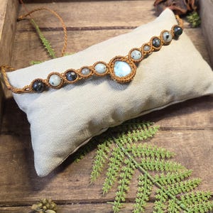 Könnte beinhalten: Ein Makramee-Armband mit einem zentralen hellblauen Stein, umgeben von kleineren runden Perlen in Grau-, Weiß- und Blautönen. Die Perlen werden von einer braunen geknoteten Kordel gehalten, mit verstellbaren Kordelenden an den Seiten des Armbands.