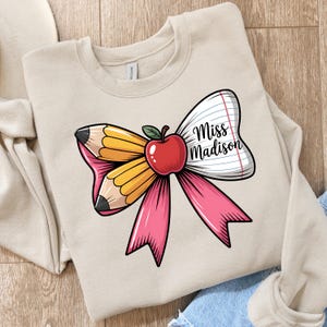 Könnte beinhalten: Beiges Sweatshirt mit einem Schleifendesign mit Bleistift, Apfel und liniertem Papier. Die Schleife ist rosa, gelb und weiß, mit dem Text "Miss Madison" auf dem Papier. Ein Design zum Thema Lehrer.