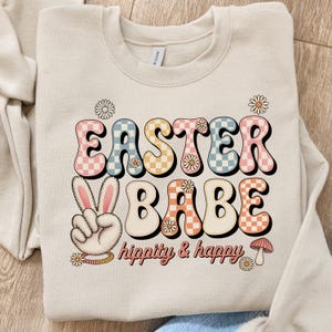 Może przedstawiać: Beżowa bluza z napisem "EASTER BABE" w stylu retro. Litery są w kratkę w kolorze różowym, niebieskim i pomarańczowym. Widoczny jest również znak pokoju z uszami królika oraz słowa "hippity & happy".