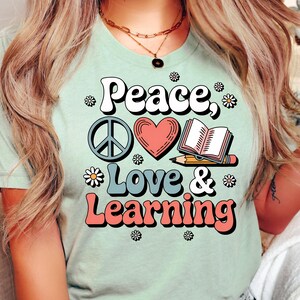 Peut inclure: T-shirt vert clair avec l'inscription "Peace, Love & Learning" dans une police rétro. Le motif comprend un symbole de paix, un cœur, un livre ouvert et des marguerites. Le t-shirt est en matière douce et confortable.