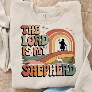 May include: Beige sweatshirt with the text "THE LORD IS MY SHEPHERD" in retro-style lettering. The design includes a rainbow, a shepherd silhouette, and a lamb on a colorful landscape. A cozy and inspirational apparel item.