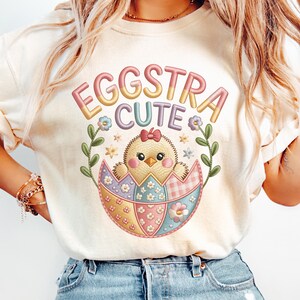 May include: A cream-coloured t-shirt with the words "EGGSTRA CUTE" in pastel shades. The design shows a chick hatching from a patchwork egg, framed by floral and leaf details. This is a casual t-shirt for everyday wear.