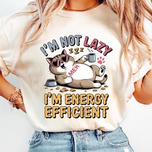 Puede incluir: Camiseta color crema con un gato de dibujos animados con gafas de sol en forma de corazón y una taza de café. El texto dice "I'M NOT LAZY, I'M ENERGY EFFICIENT". También se ve un cuenco de comida para gatos y huellas de patas.