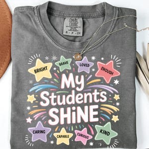 May include: A gray Comfort Colors t-shirt with the text "My Students Shine" in colorful lettering. The shirt features star-shaped graphics with words like "Bright," "Brave," and "Kind." A gold necklace with a pendant rests on the shirt.