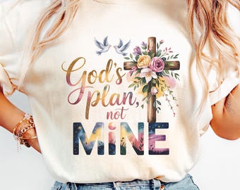 May include: Cream-colored t-shirt featuring the text "God's plan, not mine" in a colorful, script font. A wooden cross adorned with flowers and two doves is centered above the text. The shirt has short sleeves and a crew neck.