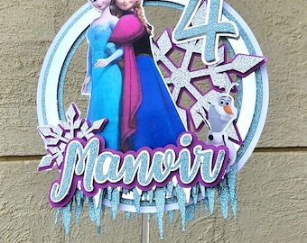 May include: A Frozen themed cake topper with Elsa and Anna, a snowflake, and the number 4. The topper also includes the name "Manovir" and a blue and white icicle design.