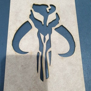 May include: A wooden stencil with a cutout of the Mandalorian symbol. The symbol is a stylized skull with horns and a vertical line down the center.