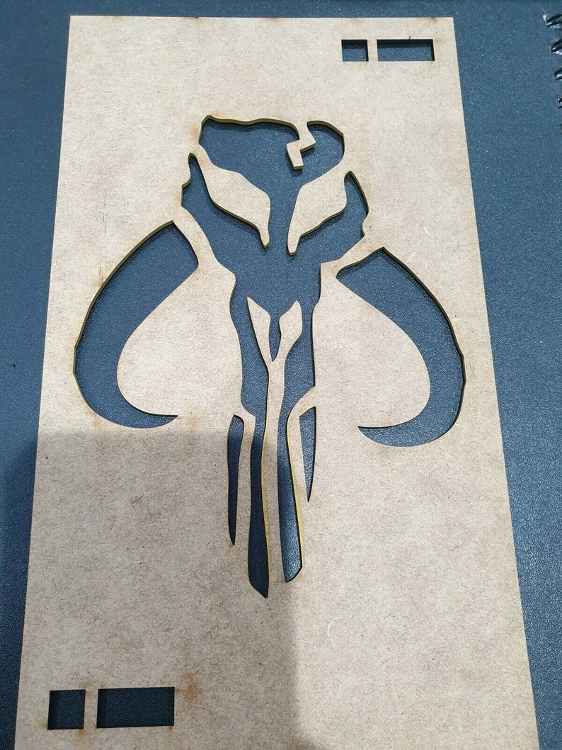 May include: A wooden stencil with a cutout of the Mandalorian symbol. The symbol is a stylized skull with horns and a vertical line down the center.