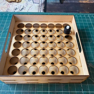 May include: A wooden paint organizer with 49 individual compartments for holding small paint bottles. The organizer has a handle on the side and is made of light brown wood. A black paint bottle is sitting in one of the compartments.