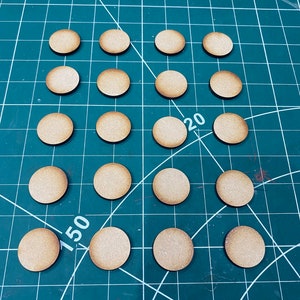 May include: A collection of 18 round wooden bases, perfect for miniature figures or game pieces. The bases are arranged in a grid pattern on a green cutting mat with white markings.
