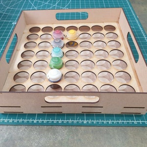 May include: A wooden paint pot holder with 48 round holes for storing paint pots. The holder has two handles on the sides and is made of light brown wood. There are several paint pots in the holder, including green, yellow, and red.