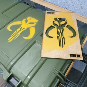 May include: A yellow stencil of the Mandalorian symbol on a wooden board. The stencil is being used to create a similar design on a green box.