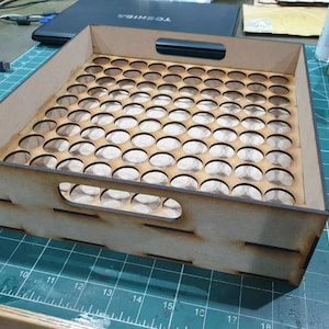 May include: A wooden tray with a grid of circular cutouts, designed to hold small round objects like paint tubes or vials. The tray has a handle on the top and is made of light brown wood.