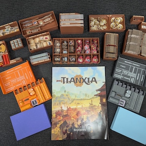 Può includere: Un organizer per giochi da tavolo con vari vassoi di stoccaggio marroni, carte e pezzi di gioco. Il tabellone di gioco intitolato "Tianxia" è visibile, insieme a componenti arancioni e blu. I componenti sono disposti su una superficie scura.
