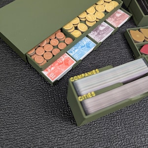 May include: An olive green game organizer with compartments holding game money and cards. The money includes brown, yellow, and gold coins, and various denominations of paper currency. The card holder has the words "COMPANIES" and "CITIES" printed on it.