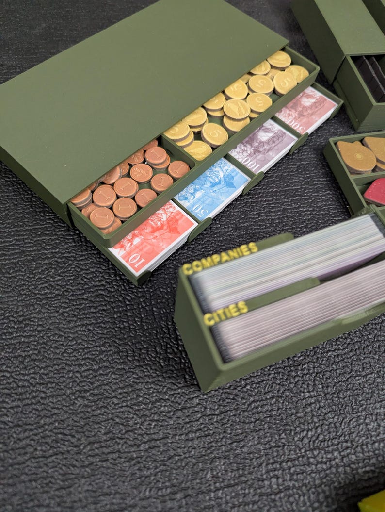 May include: An olive green game organizer with compartments holding game money and cards. The money includes brown, yellow, and gold coins, and various denominations of paper currency. The card holder has the words "COMPANIES" and "CITIES" printed on it.