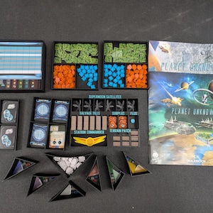 May include: A board game set, "Planet Unknown", with various components. Includes game cards, colored tokens, terrain tiles, and a rulebook. The box art features a space scene with planets and spacecraft. The game is set on a dark surface.