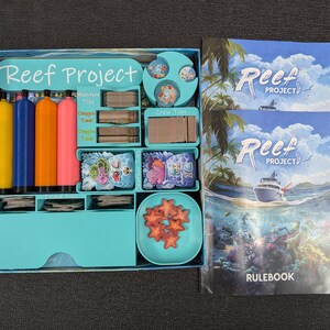 Op de afbeelding: Een turquoise blauwe gamebox met de titel "Reef Project" op de deksel. De doos bevat verschillende spelonderdelen, waaronder kleurrijke markers, dobbelstenen en tegels. De doos is open en laat de inhoud zien. Twee regelboeken met de titel "Reef Project" zijn ook zichtbaar.