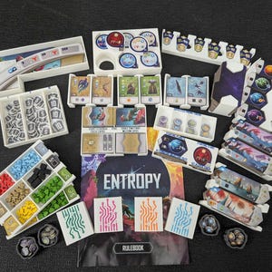 May include: A board game organizer set for Entropy, featuring white plastic trays containing various game components including colored wooden resource cubes, hexagonal tokens, card decks, player boards, and a rulebook with the title Entropy printed on the cover.