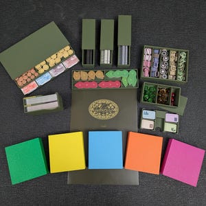 May include: An assortment of board game components, including wooden tokens, cards, and dice, arranged in olive green boxes and trays. Colorful foam squares in green, yellow, blue, orange, and pink are also present. The game appears to be themed around nature.