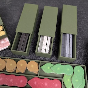 May include: An overhead shot of a board game with various components. Three olive green boxes are open, revealing black, white, and gray game pieces. Other game pieces are in a tray, including tan, red, and green shapes.