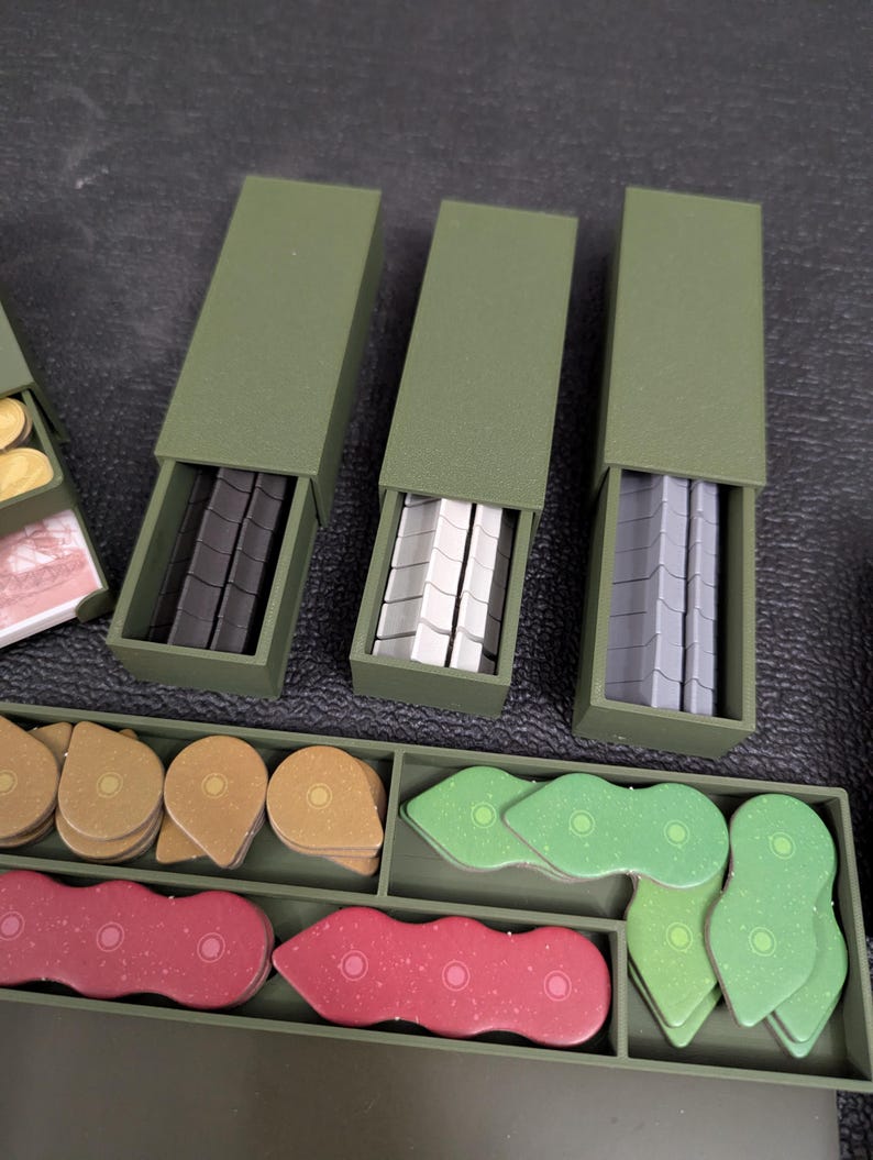May include: An overhead shot of a board game with various components. Three olive green boxes are open, revealing black, white, and gray game pieces. Other game pieces are in a tray, including tan, red, and green shapes.