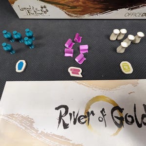 May include: A game board with the title "River of Gold" printed on it. There are several small wooden pieces in different colours and shapes on the board, including blue, pink, and brown.