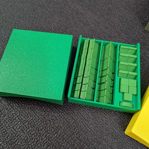 May include: A green plastic organizer with a lid, filled with various green rectangular and square pieces. The pieces are arranged in compartments, suggesting a game or organizational system. The lid is to the left, and a yellow piece is in the bottom right corner.