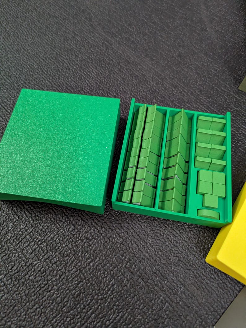May include: A green plastic organizer with a lid, filled with various green rectangular and square pieces. The pieces are arranged in compartments, suggesting a game or organizational system. The lid is to the left, and a yellow piece is in the bottom right corner.