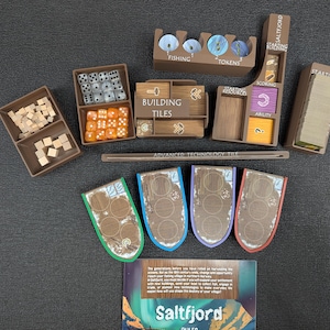 Può includere: Un set da gioco da tavolo Saltfjord con organizer in plastica marrone, cubi di legno, dadi arancioni e bianchi, quattro plance giocatore a forma di scudo con bordi verdi, blu, viola e rossi e un regolamento con un'immagine di montagne e aurora.