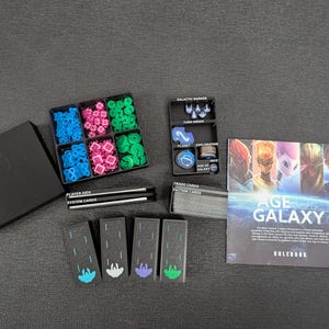 Puede incluir: Un juego de mesa, "Age of Galaxy", con varios componentes. Incluye una caja negra, un contenedor con piezas de juego azules, rosas y verdes, cartas y un libro de reglas. El libro de reglas presenta ilustraciones de personajes y el título del juego.