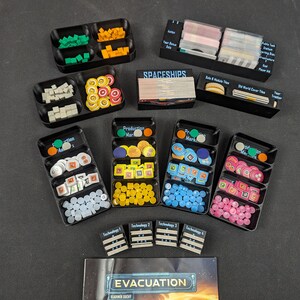 May include: A set of black plastic trays with various game pieces and tokens. The trays are labeled with the names of the game components, such as "Spaceships", "Production Markers", and "Technology 1". The game board is visible in the background.