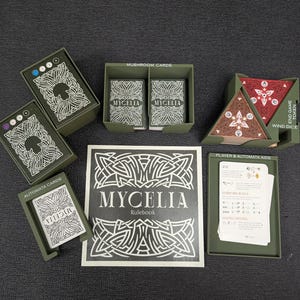 Puede incluir: Un juego de mesa con cartas, dados y un libro de reglas. El juego presenta una combinación de colores verde y blanco con la palabra "MYCELIA". El juego incluye "Mushroom Cards", "Automata Cards" y "Wind Dice".