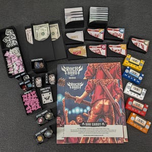 Puede incluir: Un juego de mesa titulado Rock Hard 1977 que incluye un libro de reglas, mazos de cartas, organizadores de plástico negro, fichas de cohete rosas, fichas de lazo blancas y cinco piezas de juego con forma de casete en amarillo, azul, blanco, rojo y naranja.