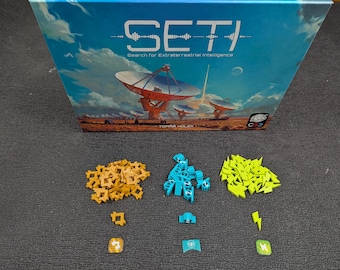 Könnte beinhalten: Ein Brettspiel namens SETI: Search for Extraterrestrial Intelligence. Die Spielbox zeigt eine farbenfrohe Illustration eines großen Radioteleskops. Das Spiel enthält eine Vielzahl von bunten Spielsteinen, darunter gelbe, blaue und grüne.