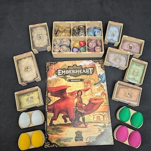 Puede incluir: Juego de mesa "Emberheart" con un libro de reglas que muestra un dragón y una persona. Incluye portatarjetas, fichas y piezas con forma de huevo de dragón en amarillo, blanco, verde y rosa. Los componentes del juego son beige.